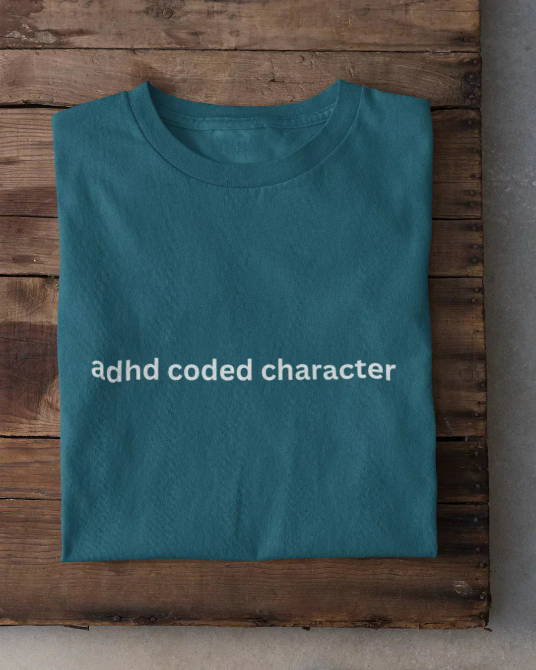 Adhd Coded Character T-Shirt in teal with white lowercase text across the front, folded on a wooden surface.