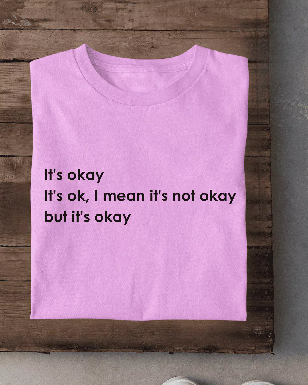 It's Okay T-Shirt - pink regular tee with black printed text, folded on wood background