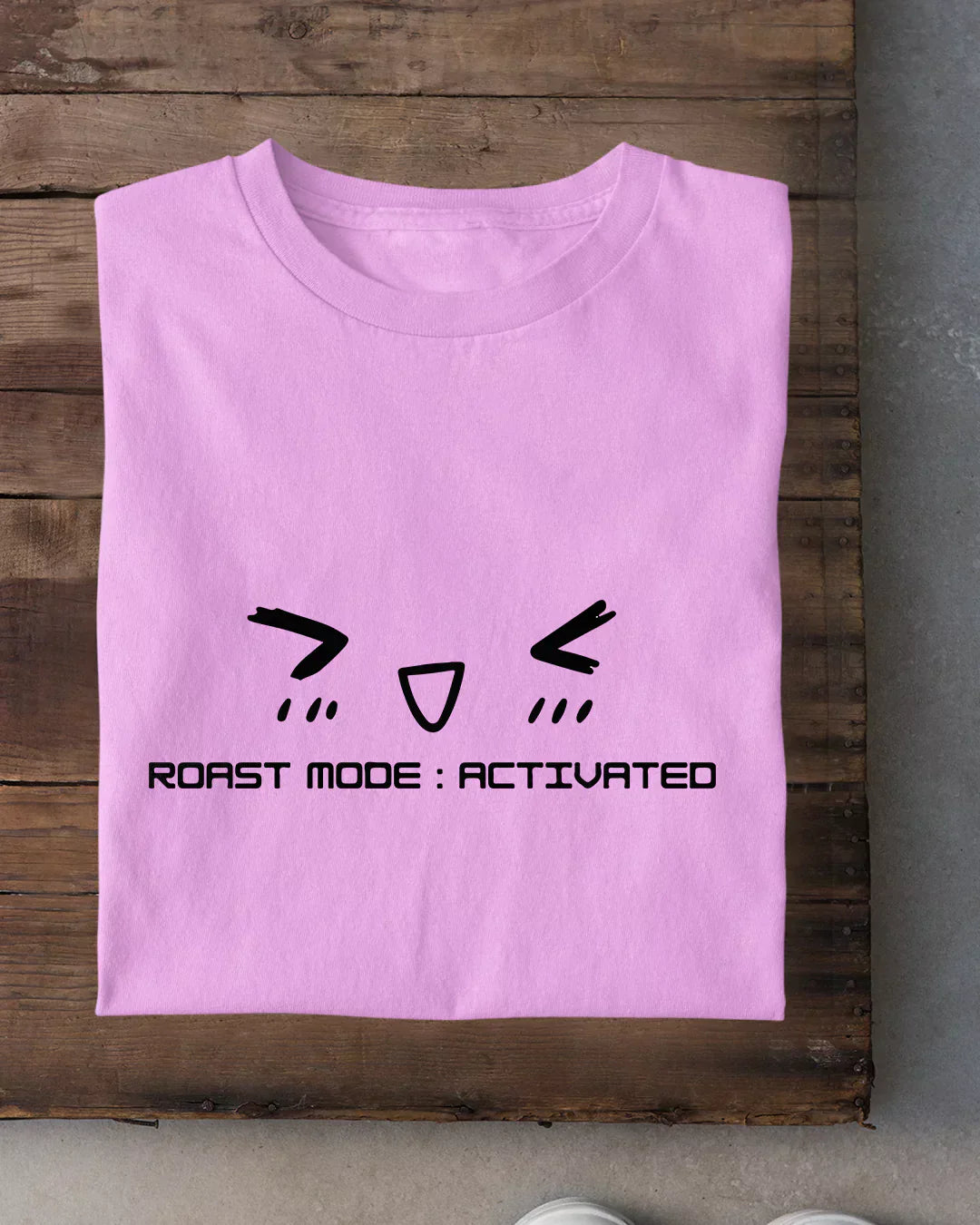 Roast Mode T-Shirt - pink regular tee with a black cartoon face graphic and ROAST MODE: ACTIVATED text, shown flat on wood.